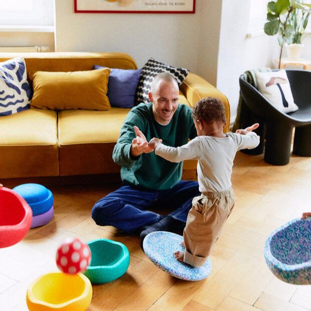 A parent helps a toddler wobble on the super cool balance board with Stapelstein stepping stones around them.