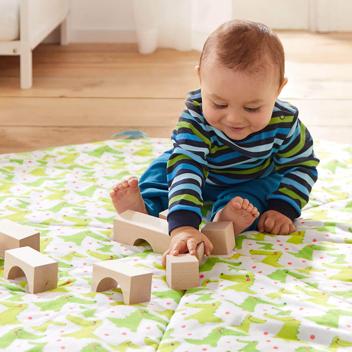 A smiling baby sits on a colorful blanket, playing with the Basic Building Blocks 26 Piece Starter Set by HABA, surrounded by a cozy indoor environment.