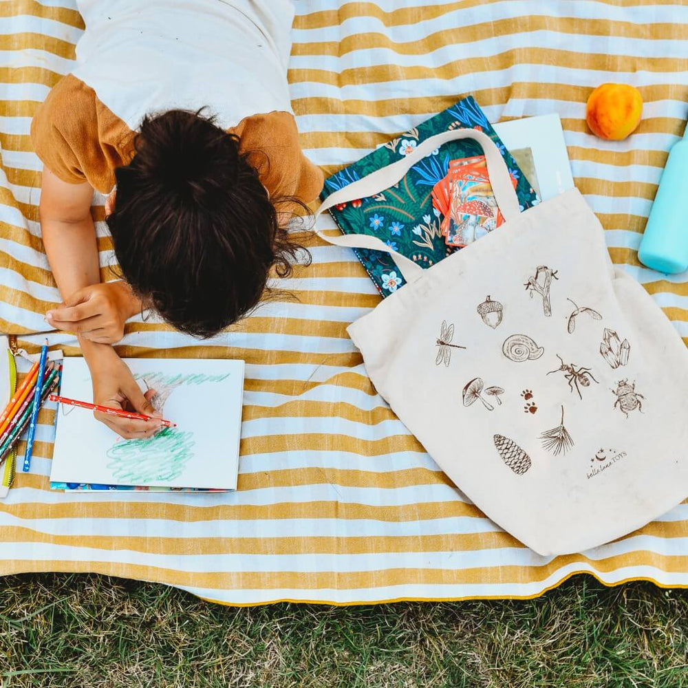 Bella Luna Toys Nature Hunt Book Tote on a picnic blanket with a child drawing.