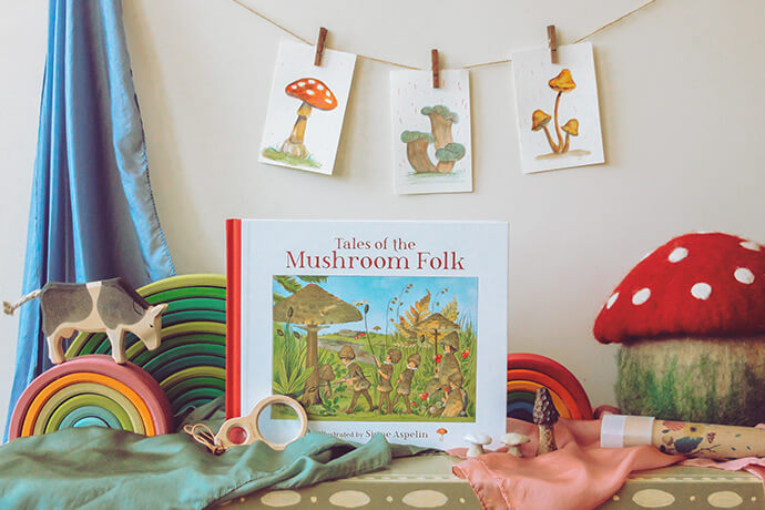 Tales of the Mushroom Folk Book on silks with mushroom paintings hanging in the background, a large stuffed mushroom with red cap, and a cow on top of a Grimm's rainbow