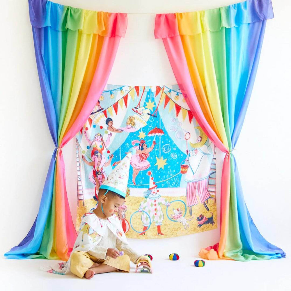 Sarah's Silks Circus Playsilk hanging on a wall with rainbow curtains and a bow in a circus silk hat playing with toys in front on the floor