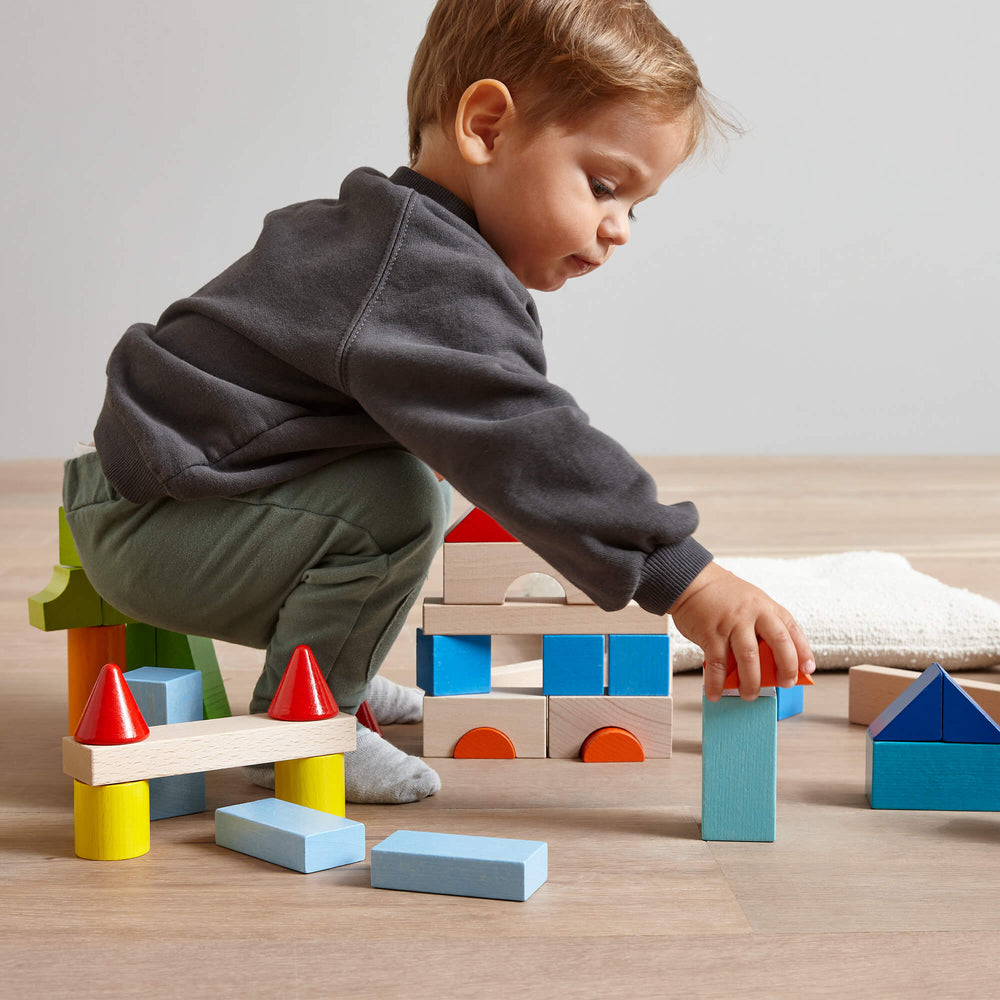 Young boy bending over and grabbing an orange wooden building block with other wooden blocks stacked in the background