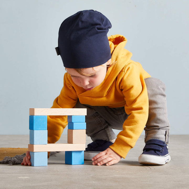 A young child in a yellow hoodie and navy beanie concentrates on building a structure with colorful blocks on the floor.