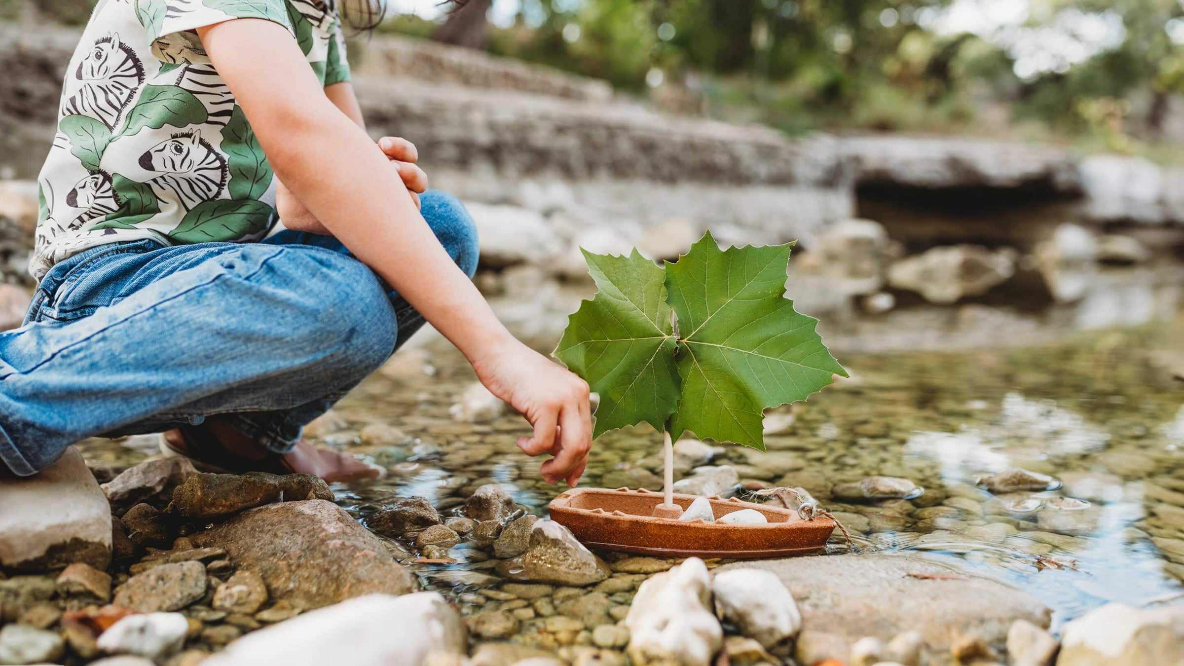 Child kneeling in stream with Cork Boat in the water with green leaf as the sail
