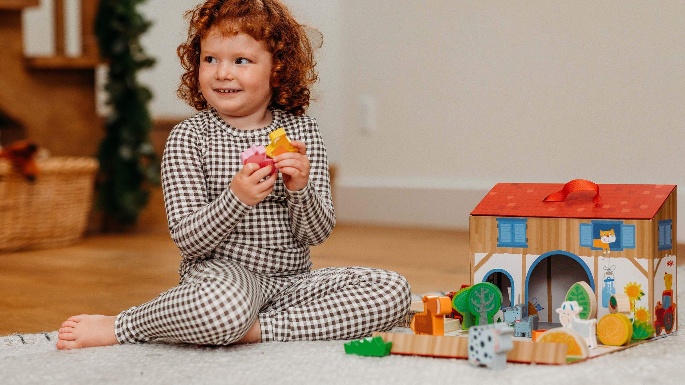 Child in checkered pajamas sitting on the floor with 2 wooden animals in her hand and HABA's Play World on the Farm next to her