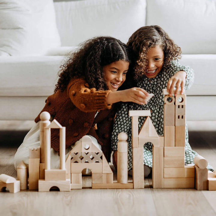 Two girls with curly hair are joyfully playing with the Basic Building Blocks 102 Piece Extra Large Wooden Starter Set, building structures on a floor, smiling as they create.