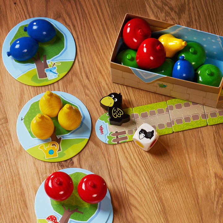 My Very First Games - First Orchard wooden fruit pieces and raven are arranged on playful game boards, alongside a small box and dice, on a wooden table