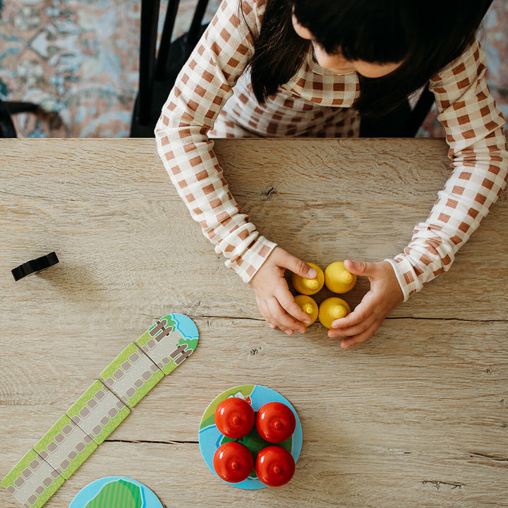 A child with dark hair holds yellow wooden pears in both hands, surrounded by wooden apples and First Orchard game pieces on a wooden table.