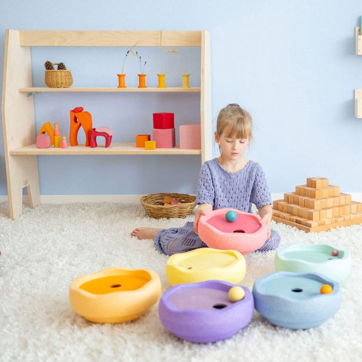 A child in a purple dress holds a Stapelstein stepping stone with pastel rainbow rolling disc. They are balancing a ball on the top. Other Grimm's and Stapelstein toys surround them.