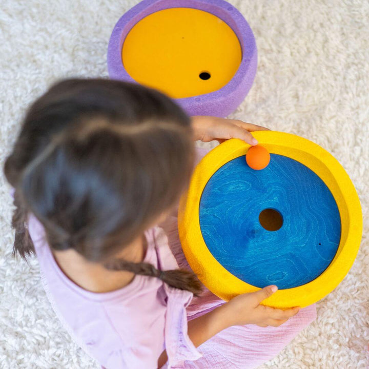 A child holding a yellow Stapelstein stepping stone in their hands. Inside is a blue Grimm's rolling disc and wooden ball.