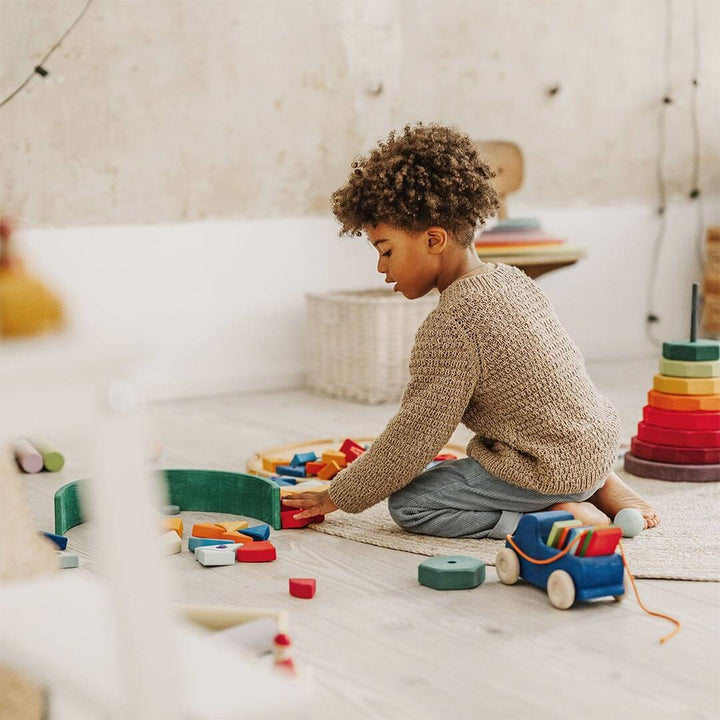 Boy playing on a playroom floor with Grimm's wooden blocks and Grimm's Wooden Pull Along Truck