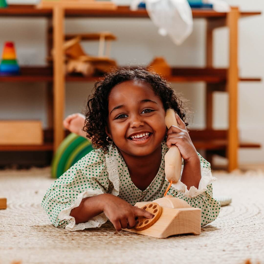 A child laying on the playroom floor dials a number on the Grimm's wooden telephone.