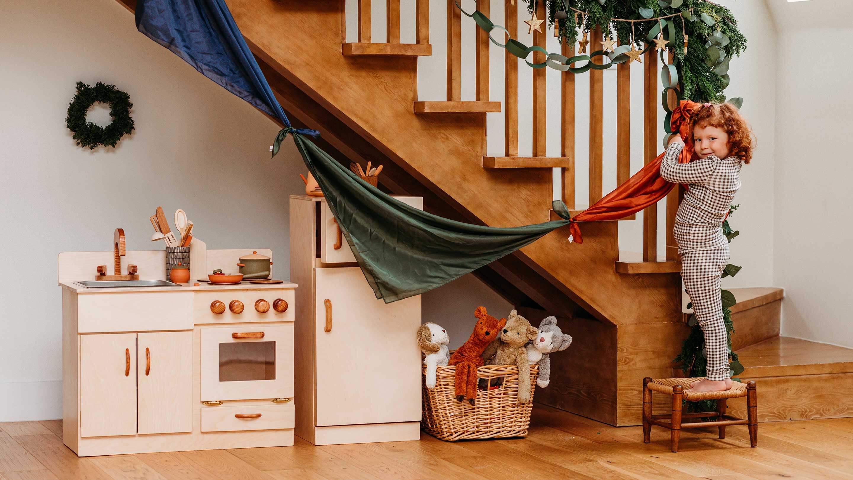 Girl holding up a string of play silks in blue, green, and red in front of a staircase with holiday decor