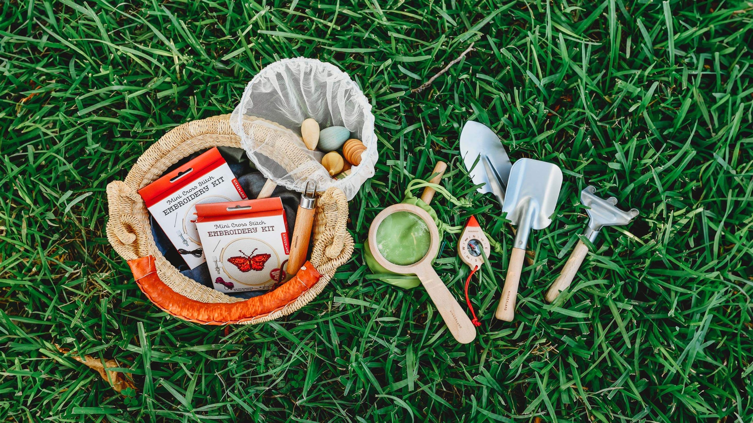 Kikkerland Embroidery Kits in a basket with a Pop Up Net laying in the grass next to a magnifying glass, compass, and garden tool set