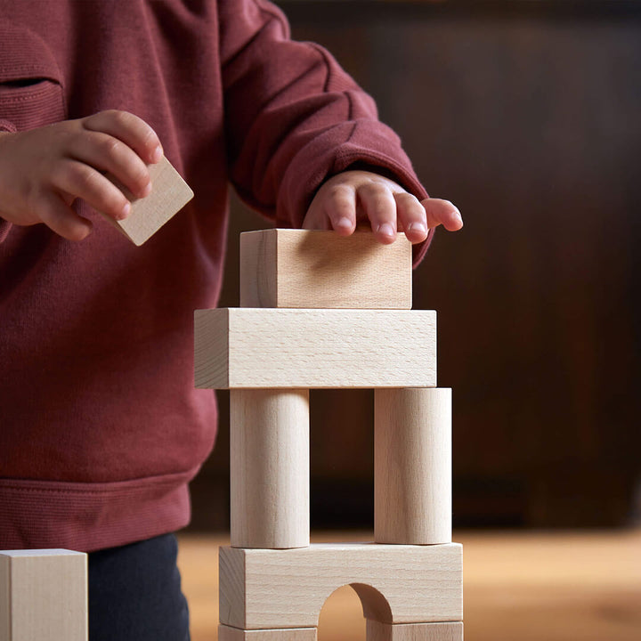 A child in a maroon sweatshirt carefully balances a wooden block on top of a stack of blocks from the Basic Building Blocks 60 Piece Large Starter Set