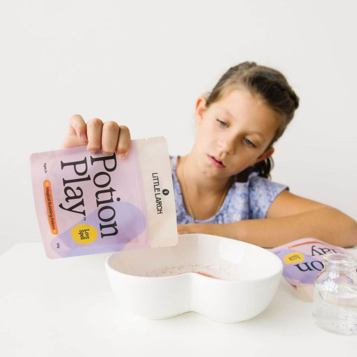 A child in a blue shirt is pouring the Little Larch Love Spell Potion Play mixture from the package into a white heart shaped bowl on the table in front of them.