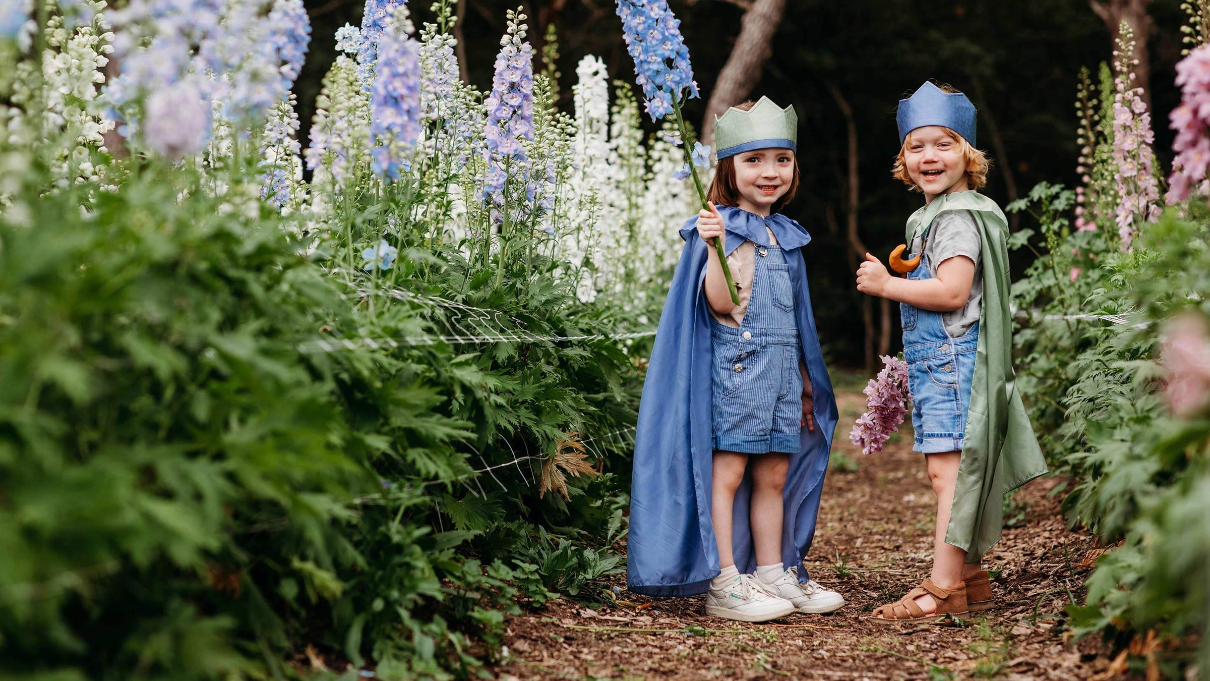 Two children wearing playsilk capes and crowns in blue and green in a field of flowers