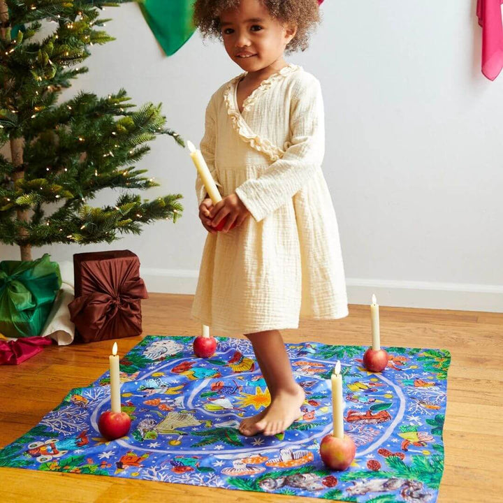 A child holding a candle walks on the Phoebe Wahl illustrated advent spiral playsilk by Sarah's Silks.