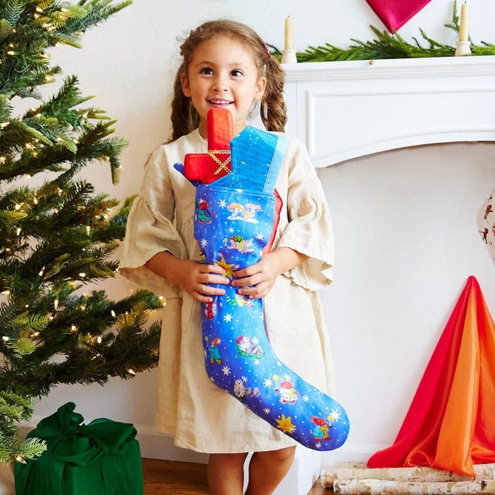 A child holds the illustrated blue silk stocking from Sarah's Silks next to a Christmas tree.