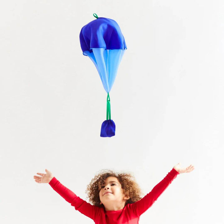 A child in red pajamas has their arms spread wide open trying to catch the falling silk blueberry parachute.