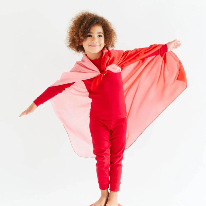 A child in red pajamas with the Strawberry Blend playsilk tied around their shoulders like a cape.