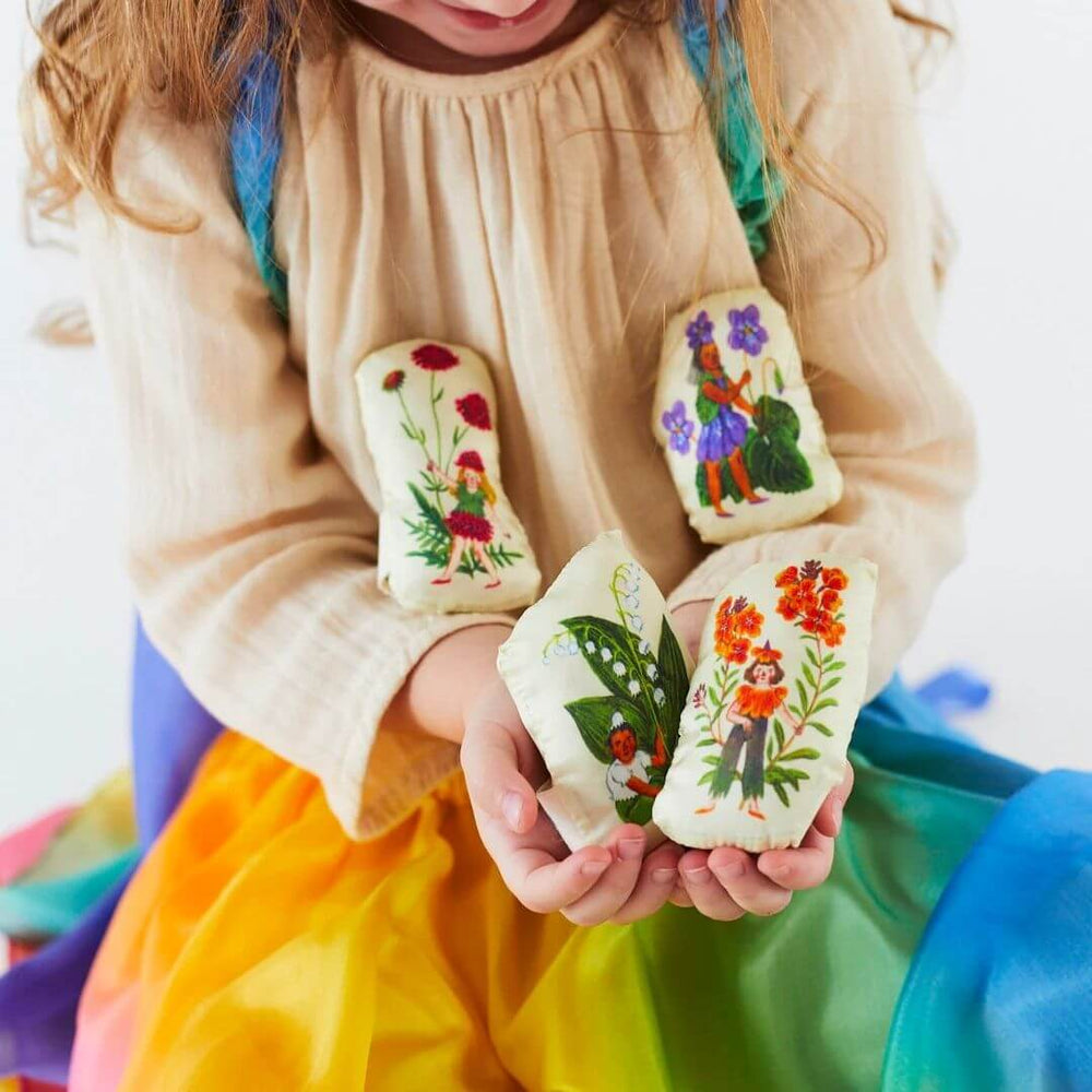 A child in a silk rainbow skirt holding all four of the Sarah's Silks Stuffed Flower Fairy figures, featuring Phoebe Wahl’s whimsical illustrations on soft silk-covered pillows.