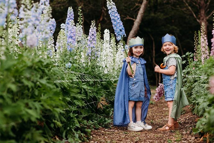 Two kids dressed in Play Silk Capes and Play Silk Crowns standing in a field of flowers